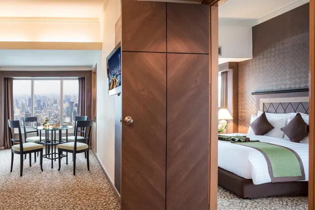 A hotel suite living area with a round dining table and chairs by floor-to-ceiling windows showing a city skyline, with a king bed visible in the adjacent bedroom.