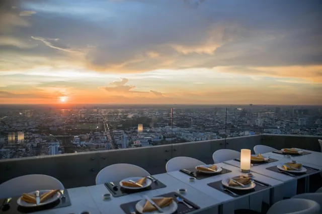 A rooftop restaurant table set with plates, folded napkins and a candle overlooks a city skyline at sunset.