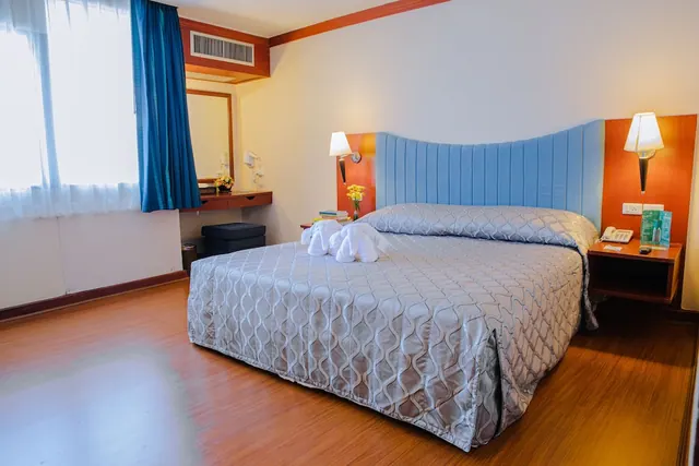 A bedroom with a large bed under a grey quilted cover and a blue padded headboard, a wall-mounted desk with a mirror, and a window with blue curtains.