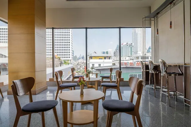 A lounge area with round wooden tables, dark-cushioned chairs, bar stools and floor-to-ceiling windows looking onto city high-rises.