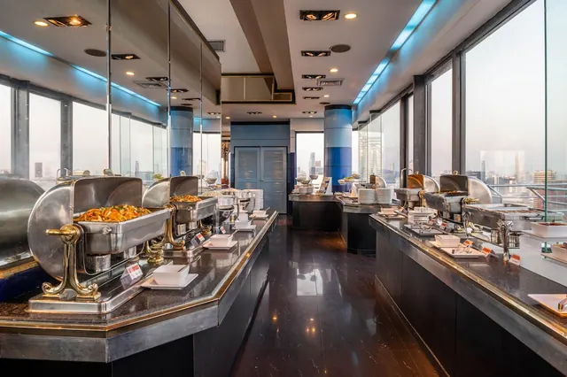 A buffet line with chafing dishes and plates runs along a restaurant with floor-to-ceiling windows overlooking the city skyline.