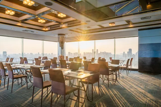 A restaurant dining room with empty tables and chairs, with floor-to-ceiling windows showing a city skyline in warm sunlight.