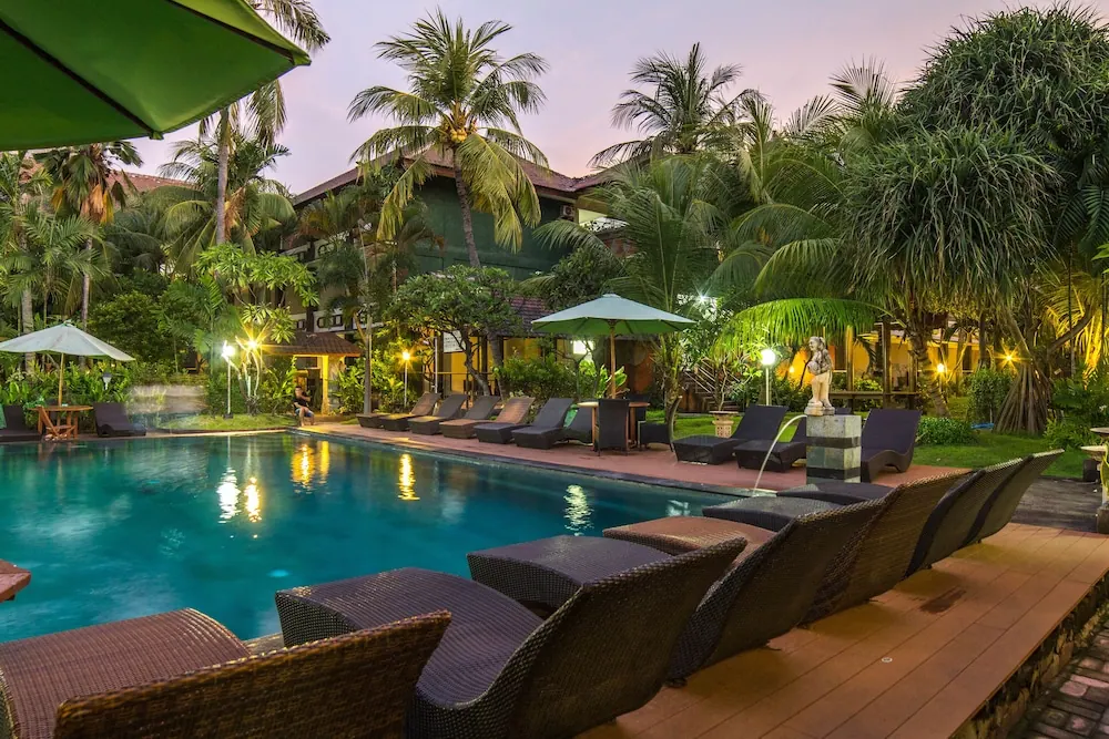 Illuminated swimming pool at dusk with wicker sun loungers, parasol umbrellas, palm trees and a statue fountain beside the pool.