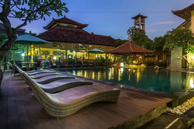 Dusk view of a pool with wicker sun loungers on a timber deck, teal umbrellas, and lit traditional Balinese pavilions behind.
