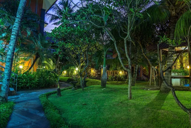 A lawn, frangipani and palm trees, and a stone path are lit by garden lights at dusk, with a lit building visible in the background.