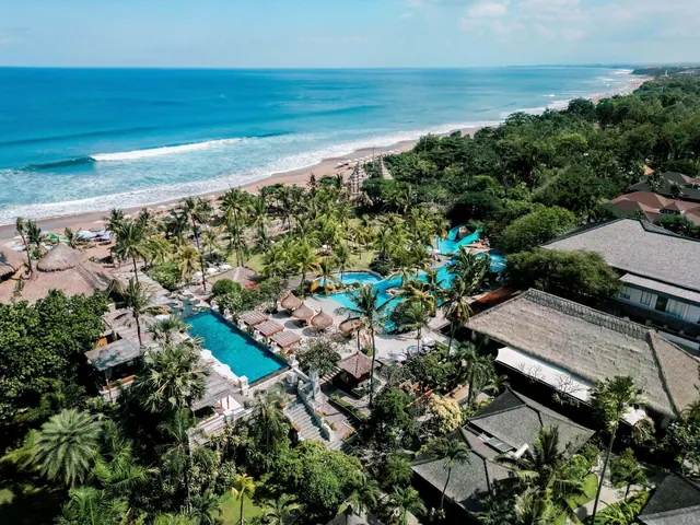 Aerial view of a beachfront resort with a rectangular pool, a curved blue pool, palm trees, thatched-roof buildings and the ocean beyond.