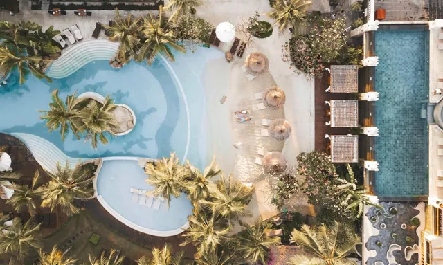 Aerial view of a freeform swimming pool beside a sandy lounge area with thatched umbrellas, sun loungers and palm trees, with a rectangular pool on the right.
