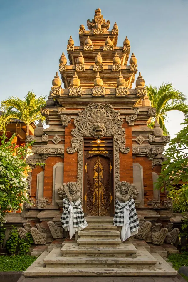 The front of a Balinese-style entrance building with carved stone ornaments, a wooden door, and two guardian statues wearing checkered sarongs, with a flight of steps leading up to it.