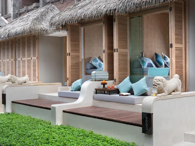 A covered terrace with cushioned daybeds, a low table holding drinks and fruit, and stone guardian lion statues beside thatched-roof buildings.