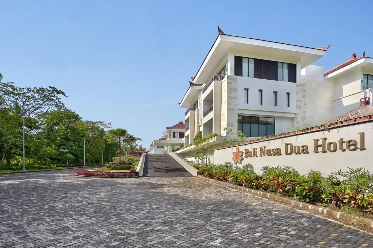 A paved driveway leads to a white multi-storey hotel building with Balinese-style roof ends, and a sign on a wall reads Bali Nusa Dua Hotel.