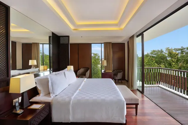 A bedroom with a made king bed, two armchairs, and sliding glass doors opening onto a balcony with a view of green trees.