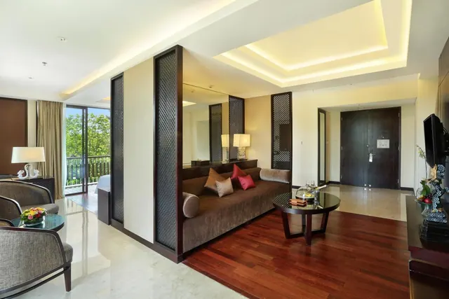 A living area with a brown sofa, a round glass coffee table, a wall-mounted television and an entrance door, with a balcony railing and trees visible through the glass doors on the left.