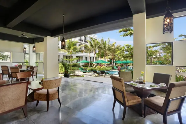 An open-air dining area with upholstered chairs and tables looks out over a pool with green umbrellas, loungers and palm trees.