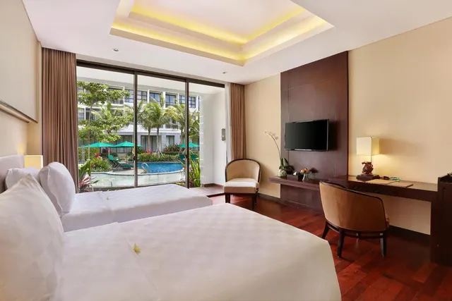 A hotel bedroom with two beds, a wall-mounted television, a desk and armchairs, with sliding glass doors looking out onto a pool and palm trees.