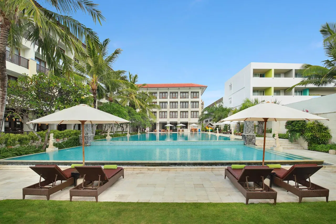 A large outdoor swimming pool with white umbrellas and brown sun loungers beside it, with hotel buildings and palm trees behind.