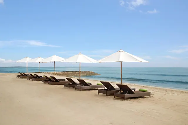 Rows of brown sun loungers and white parasol umbrellas stand on a sandy beach facing the sea under a clear blue sky.