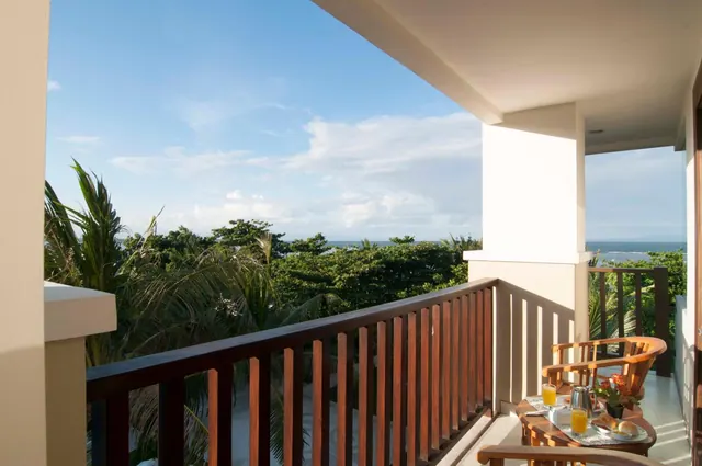 A covered balcony with a wooden railing and a small table set with juice and bread, overlooking palm trees with the sea visible on the horizon.