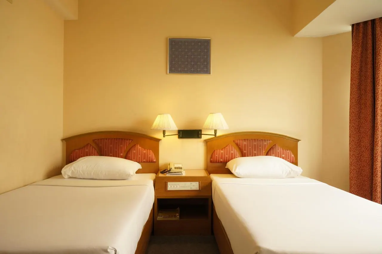 Two single beds with white bedding and wooden headboards sit side by side in a room with warm yellow walls, two wall lamps and a telephone on the nightstand between them.
