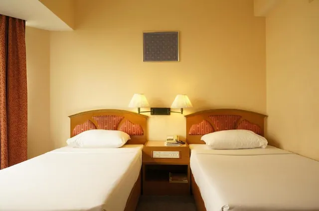 Two single beds with wooden headboards and white bedding stand either side of a nightstand with a telephone, under wall lamps.