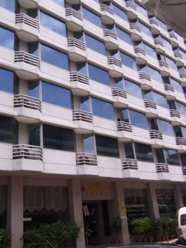 Exterior of a multi-storey building with a balconied facade, a ground-floor entrance with signage, potted plants and a white van at the right edge.