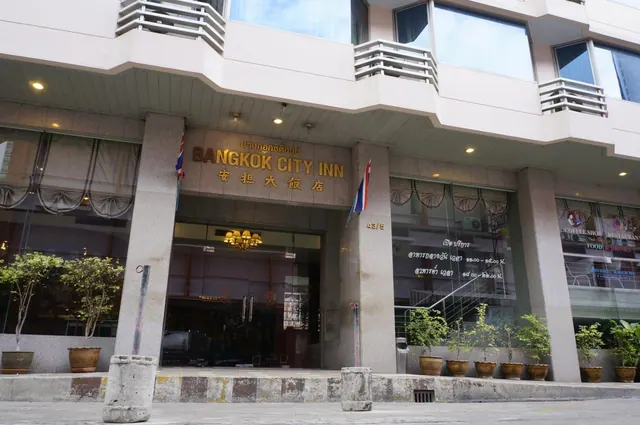 Street-level view of the Bangkok City Inn entrance with its name sign, glass doors, potted plants along the frontage and Thai flags on the columns.