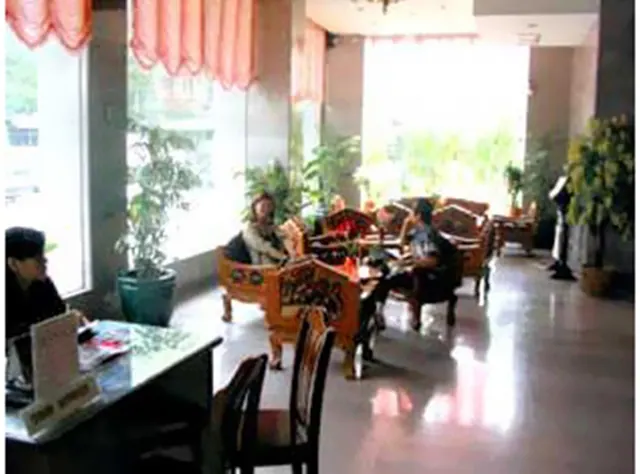 A lobby seating area with carved wooden armchairs, potted plants and a few seated people, with a reception desk at the left edge.