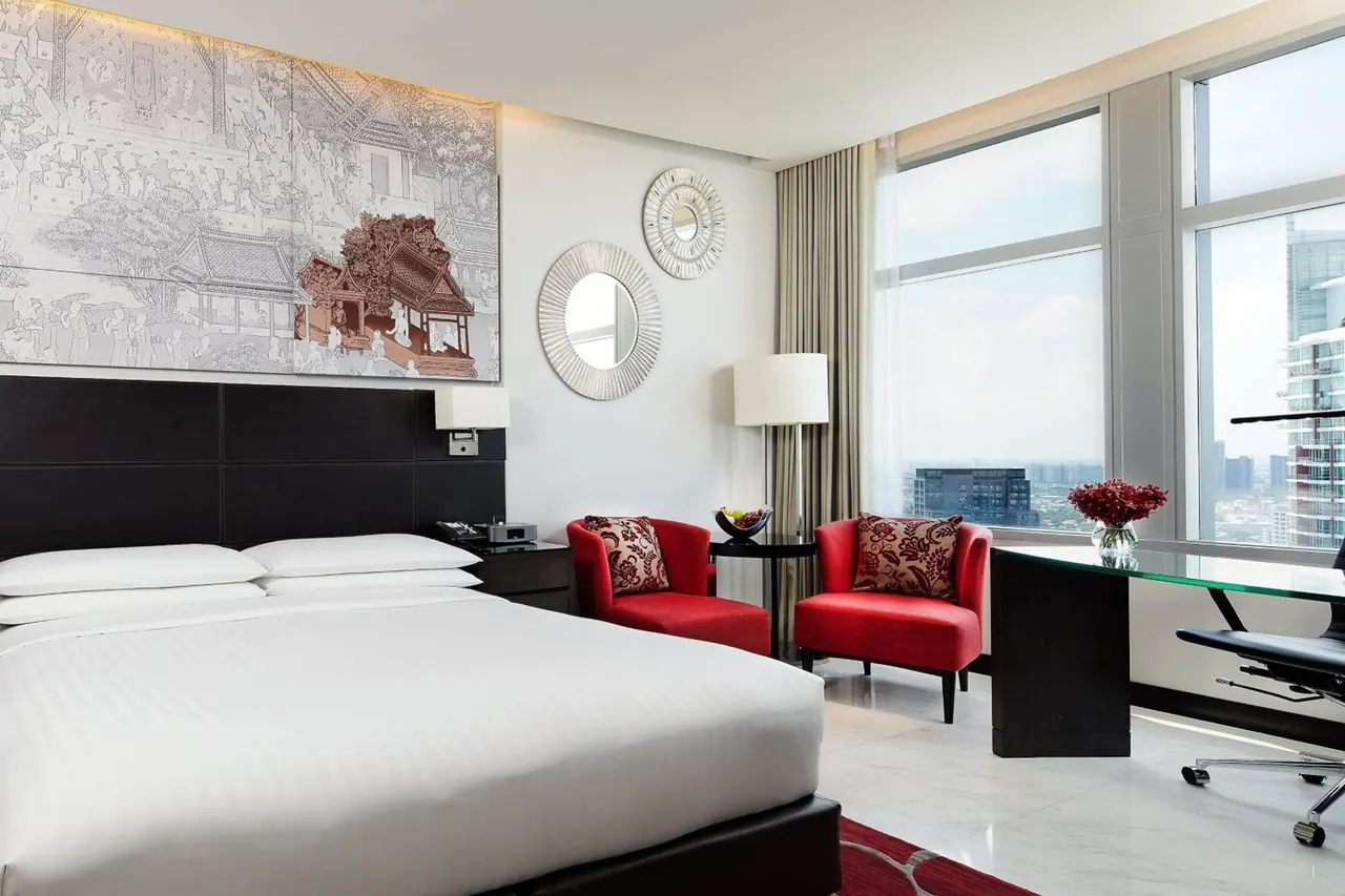 A bedroom with a king bed, two red armchairs, a glass-topped desk and floor-to-ceiling windows overlooking city buildings.