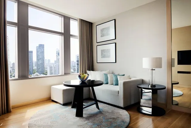 A living area with a light grey sofa, black round tables and a table lamp, beside floor-to-ceiling windows overlooking high-rise towers.