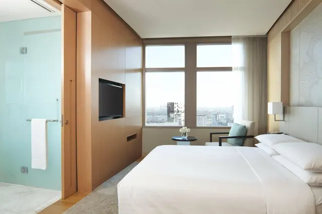 A bedroom with a king bed, a wall-mounted television, an armchair by a window, and a glass-walled shower beside the entrance, with a city skyline visible outside.