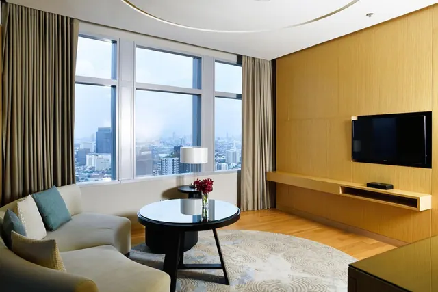 A living area with a curved sofa, round black coffee table, wall-mounted television and large windows overlooking a city skyline.