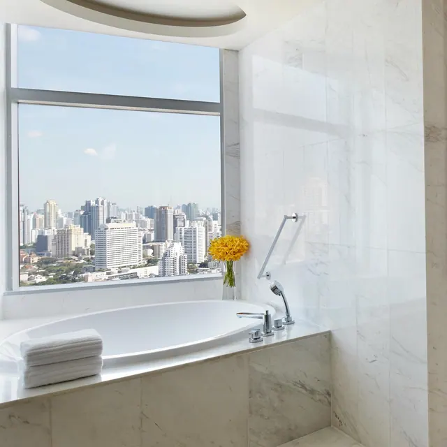 A marble bathroom with an oval bathtub, folded towels, a vase of yellow flowers and a window overlooking a city skyline of high-rise buildings.
