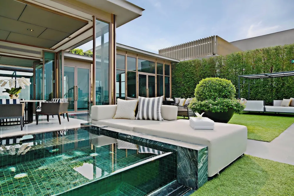 A covered terrace with a small green-tiled plunge pool, a white daybed with striped cushions, dining chairs under the roof, and a lawn beside a clipped hedge and shrub.