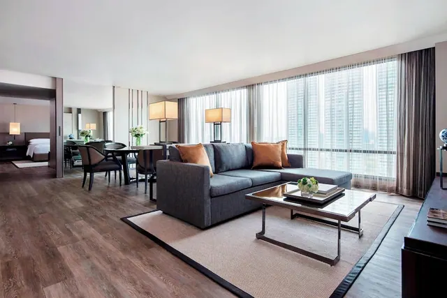 A living area with a grey sectional sofa, a glass coffee table and a dining table, with floor-to-ceiling windows showing tall buildings outside and a bedroom visible through the doorway on the left.