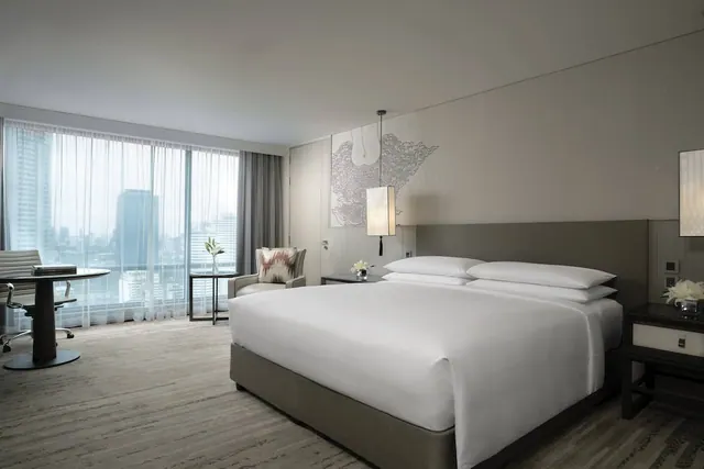 A bedroom with a king bed, an armchair and a round work desk beside floor-to-ceiling windows overlooking high-rise buildings.