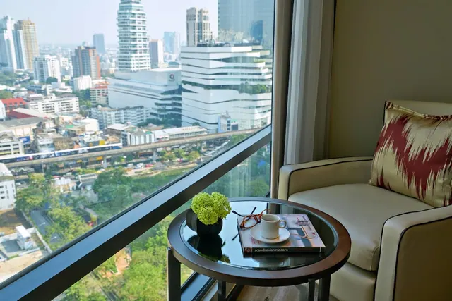 A cream armchair with a patterned cushion sits beside a round glass-top side table holding a plant, coffee cup, glasses and a book, with a floor-to-ceiling window showing city towers and an elevated train line.