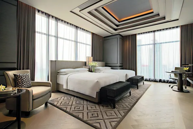 A hotel bedroom with two beds, an armchair, a desk by the window, and floor-to-ceiling windows behind sheer curtains.