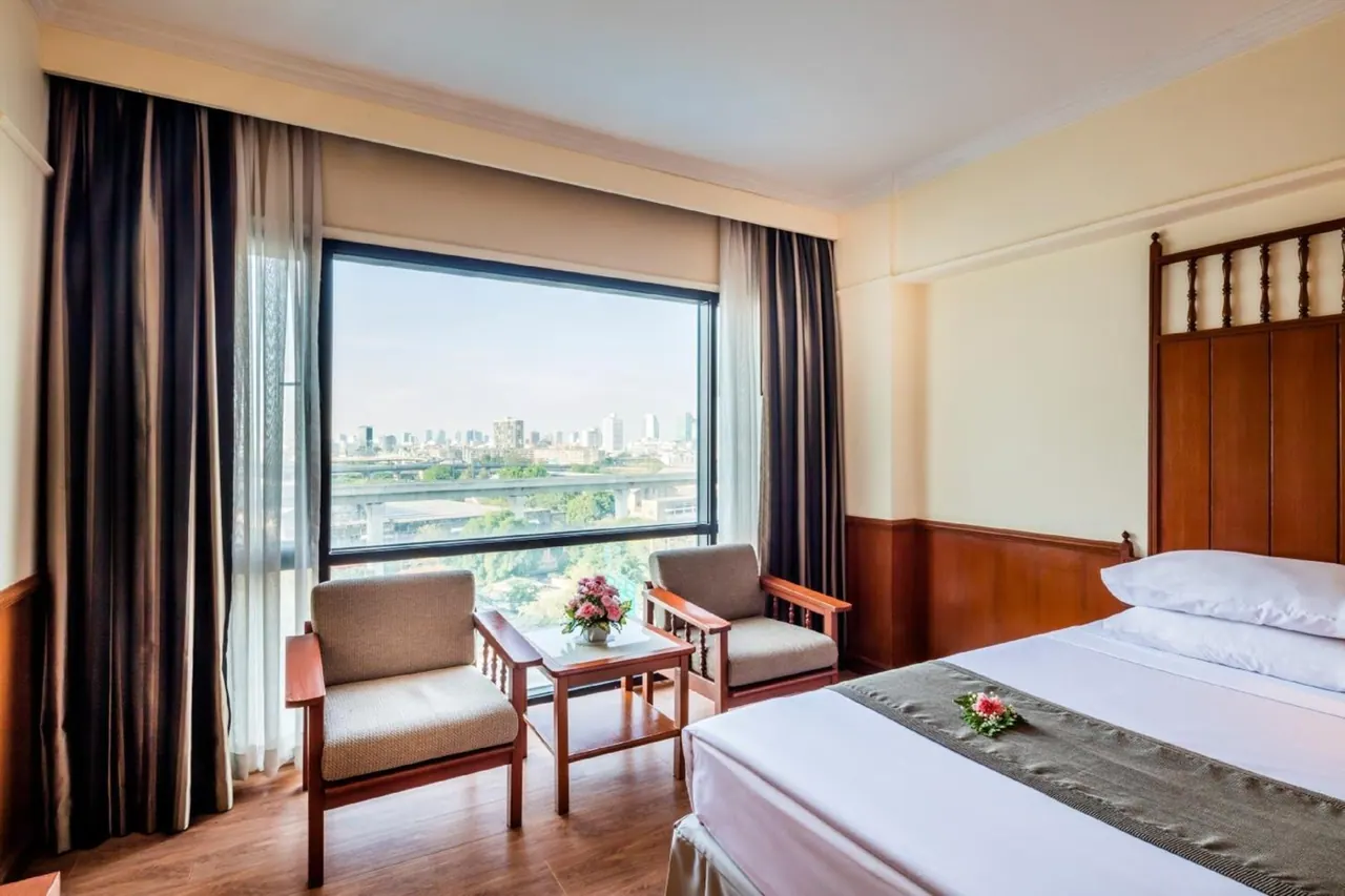 A bedroom with a double bed, two armchairs and a side table in front of a large window showing a city skyline.