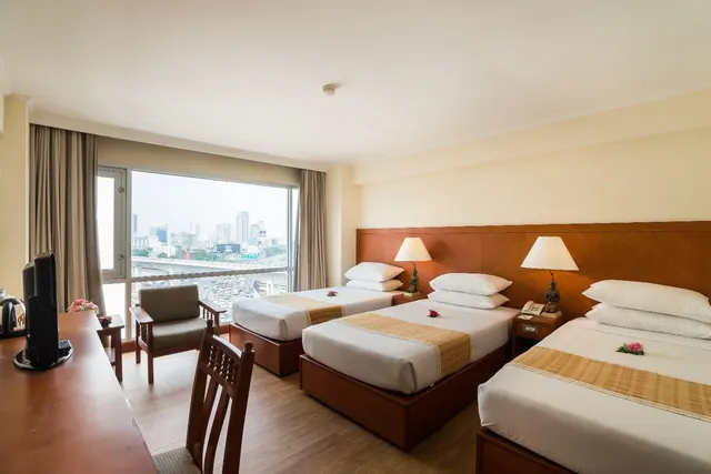 A hotel room with three beds, a desk and an armchair, and a window view over city buildings and an elevated road.