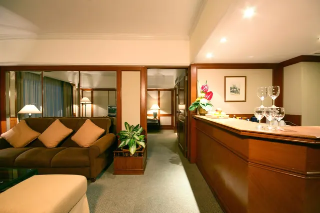 A suite living area with a brown sofa and cream ottoman, a wooden bar counter with wine glasses on the right, and a corridor leading to a bedroom in the background.