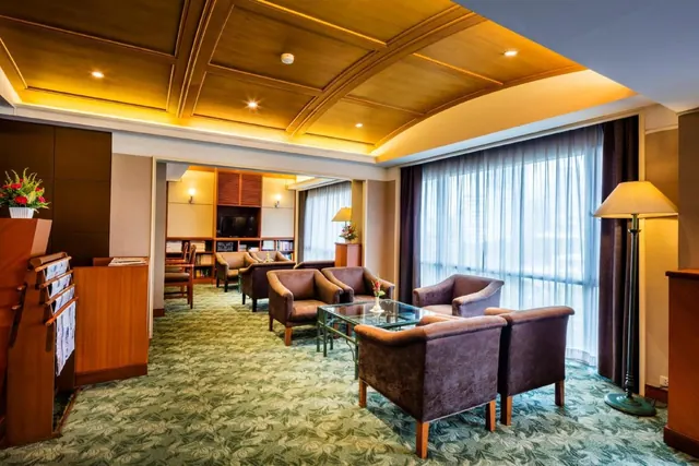 A hotel lounge with armchairs around a glass coffee table, bookshelves, a floor lamp and tall sheer-curtained windows under a wood coffered ceiling.
