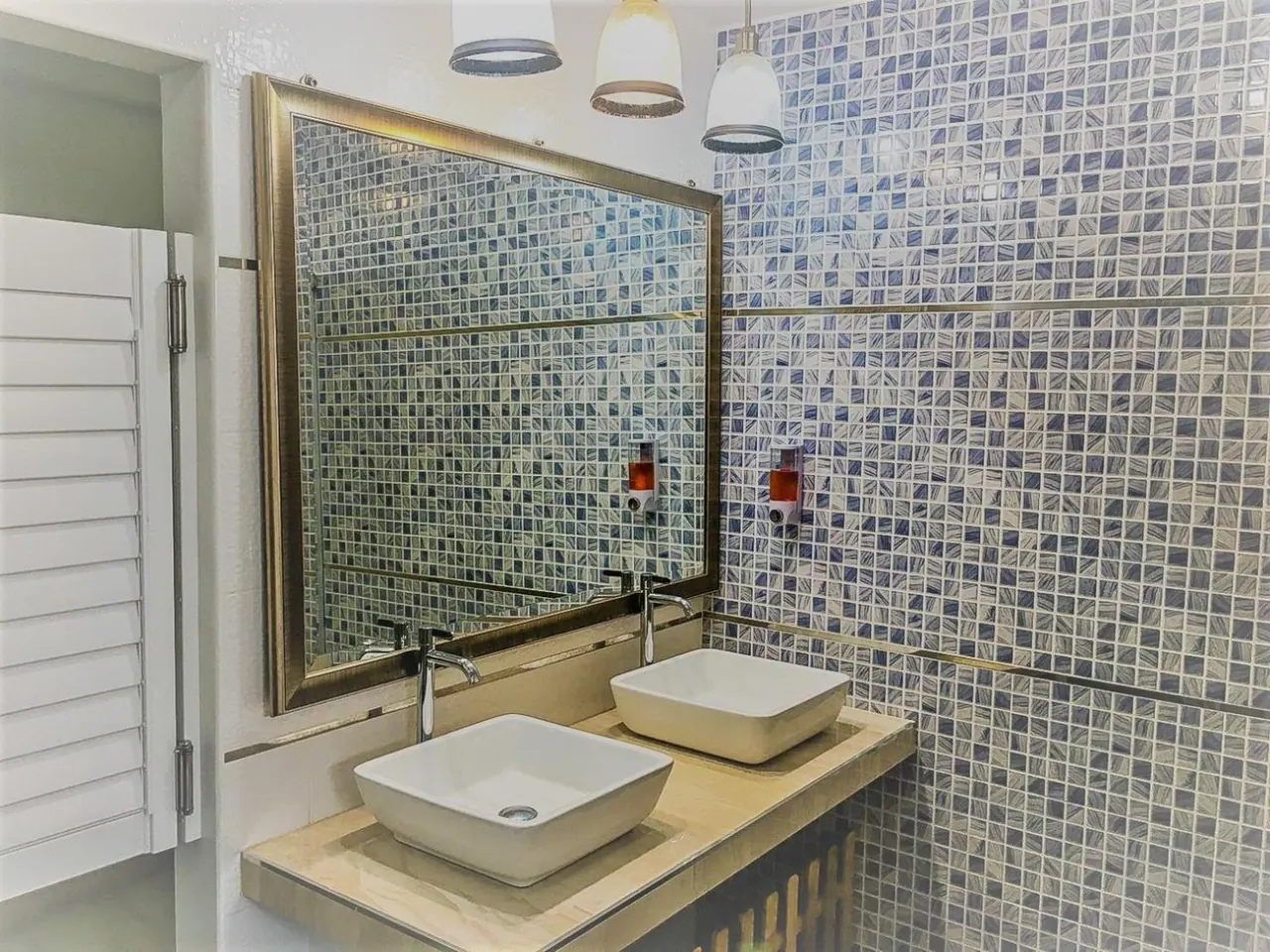 A bathroom with two white vessel sinks on a stone vanity beneath a framed mirror, with mosaic-tiled walls and pendant lights above.