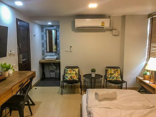 A hotel bedroom with a bed, a wooden desk and chair, two armchairs with floral cushions, a wall-mounted air conditioner, and an open bathroom with a vanity sink.