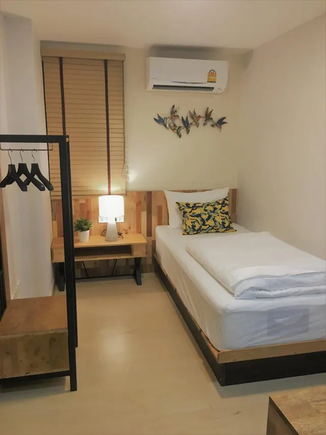 A small bedroom with a single bed, a wooden bedside table holding a lit lamp and a potted plant, a open clothes rack, and a wall-mounted air conditioner.