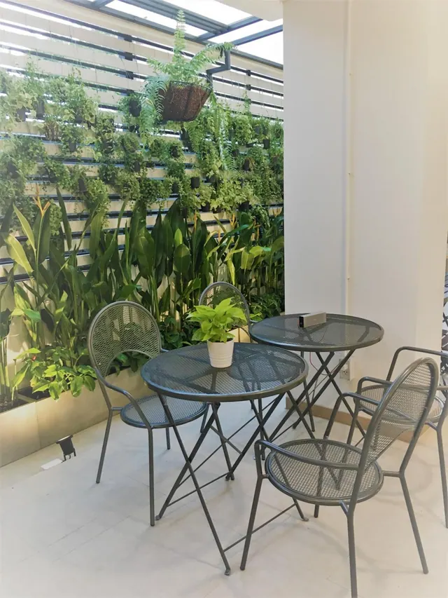Metal mesh tables and chairs sit on a tiled terrace in front of a wall of potted plants under a glass skylight.