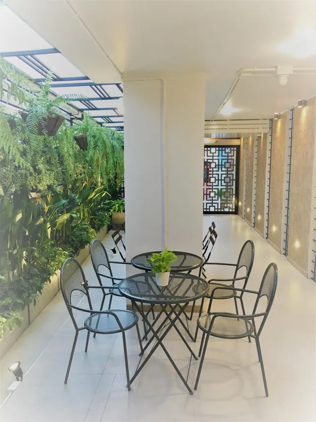 A covered terrace with metal tables and chairs beside a wall of potted plants and hanging ferns, with a lattice door in the background.