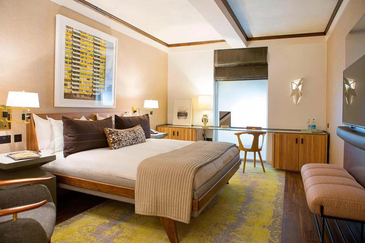 A bedroom with a king bed, a bench at the foot of the bed, a wooden desk and chair by the window, and a wall-mounted television.