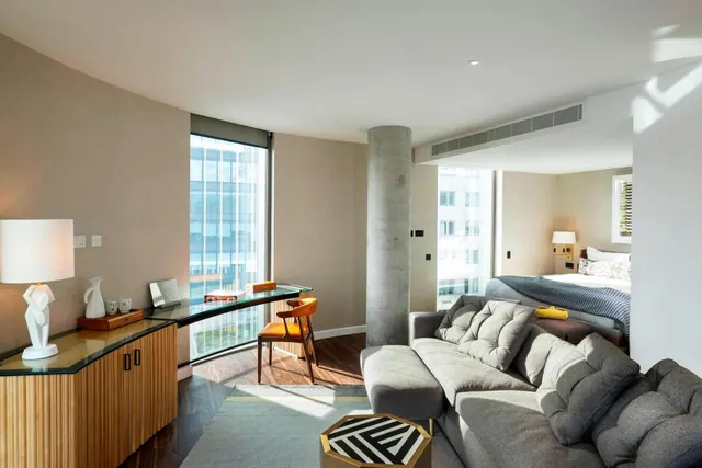 A hotel suite living area with a grey sofa, a desk with orange chairs by tall windows, and a bed visible in the background.