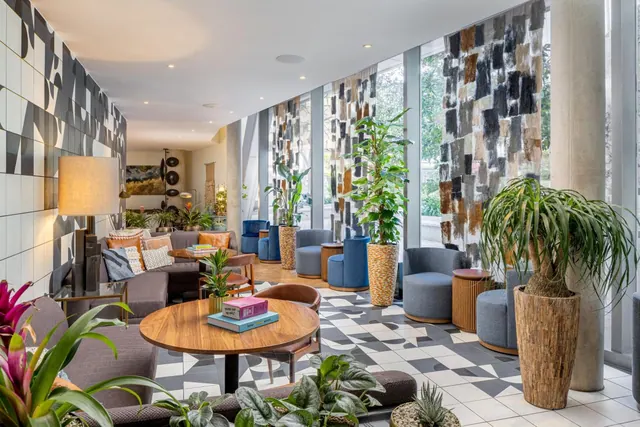 A lobby lounge with grey armchairs, sofas, a round wooden table and potted plants on a patterned tiled floor, with tall windows showing trees outside.