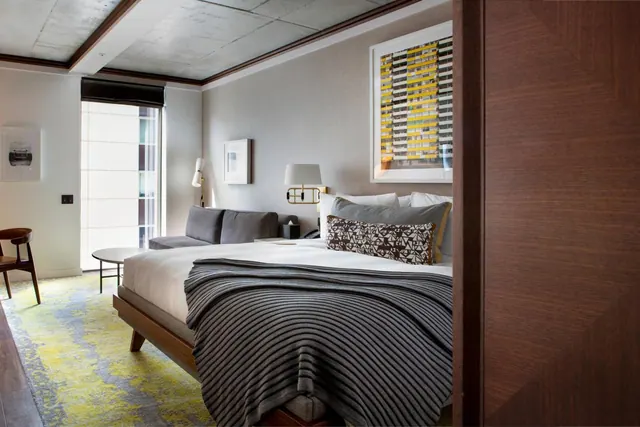 A bedroom with a made king bed, a grey sofa, a yellow patterned rug and a tall window facing a building opposite.