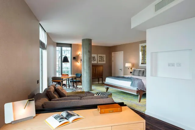 A hotel room with a grey sofa in the foreground, a round dining table by the window, a concrete column, and a double bed on the right.